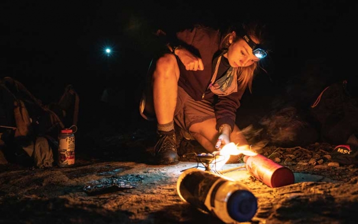 A person wearing a headlamp stokes a small fire while backpacking with outward bound. 